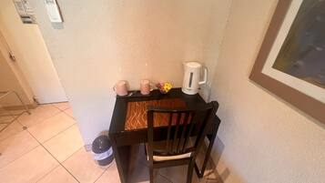 Un pequeño comedor con una mesa, dos sillas, un hervidor y un cubo de basura.