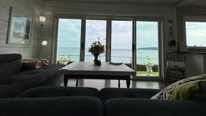 Habitación con un amplio ventanal corredero que da a un balcón con vista al mar. En el centro de la habitación hay una mesa con un jarrón de flores.