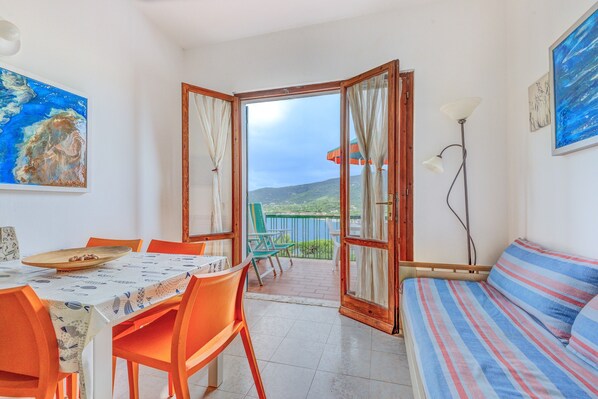 A room with a dining table, chairs, a sofa, a balcony with a view, and a painting on the wall.
