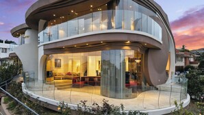 A modern, multi-story building with a curved facade, large glass windows, and a spacious outdoor seating area. - The Wave House | Custom Gem w/ Panoramic Views, Fire Pit, BBQ, Pool Table & Wraparound Balconies (San Diego)