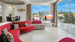 A modern living room with a red sofa, a patterned ottoman, and a dining area with a table and chairs. A TV is mounted on the wall, and a large window offers a view of the city. - The Wave House | Custom Gem w/ Panoramic Views, Fire Pit, BBQ, Pool Table & Wraparound Balconies (San Diego)