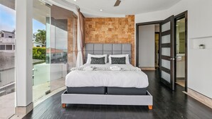 A modern bedroom with a large bed, a ceiling fan, and a view through a glass door to the outside. - The Wave House | Custom Gem w/ Panoramic Views, Fire Pit, BBQ, Pool Table & Wraparound Balconies (San Diego)