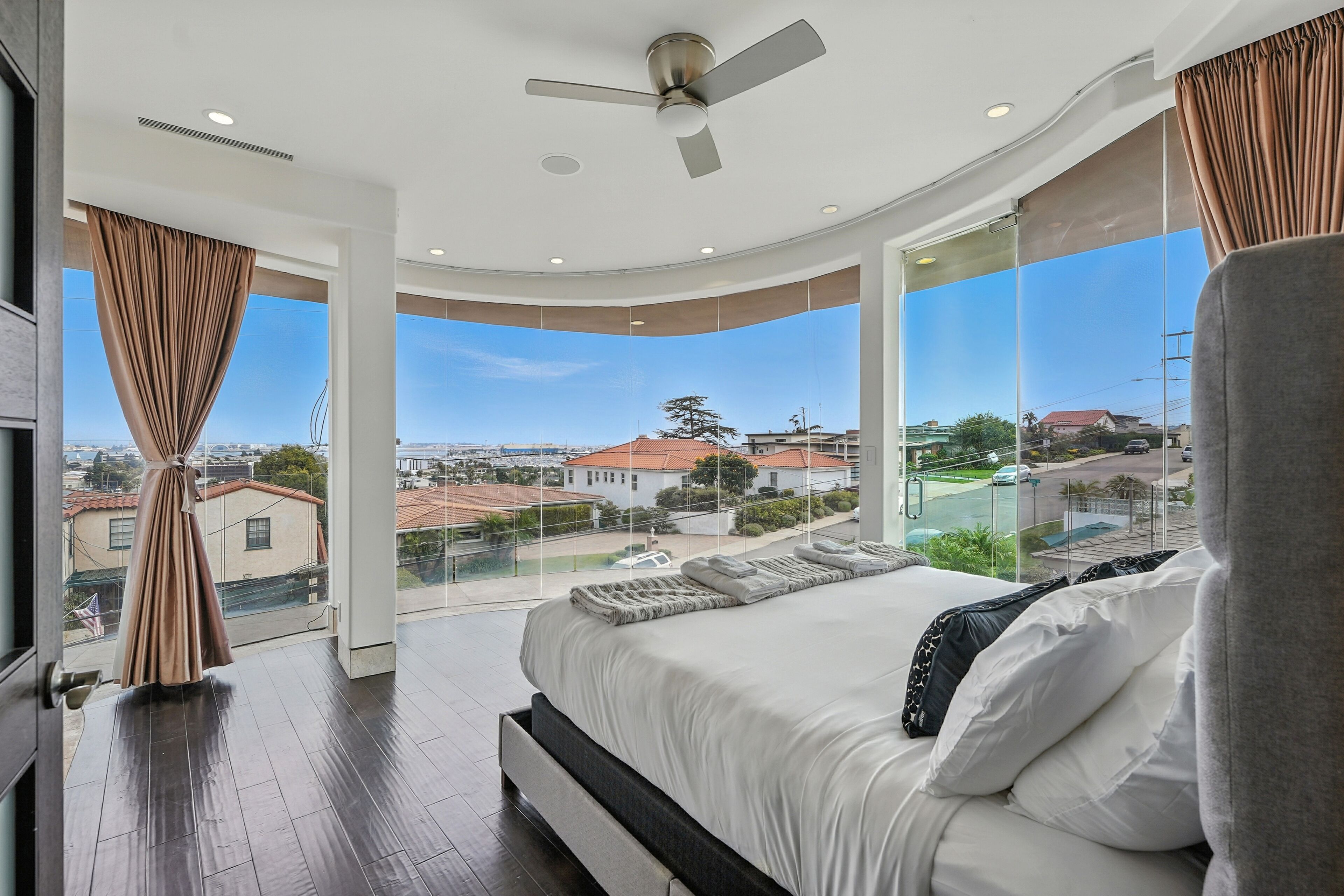 A modern bedroom with a large bed, a ceiling fan, and a view of the city through large windows.
