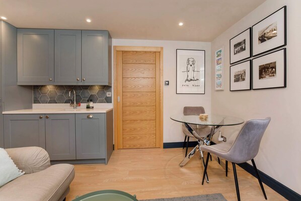 A compact kitchen with grey cabinets, a wooden door, and a dining area with a glass table and grey chairs.