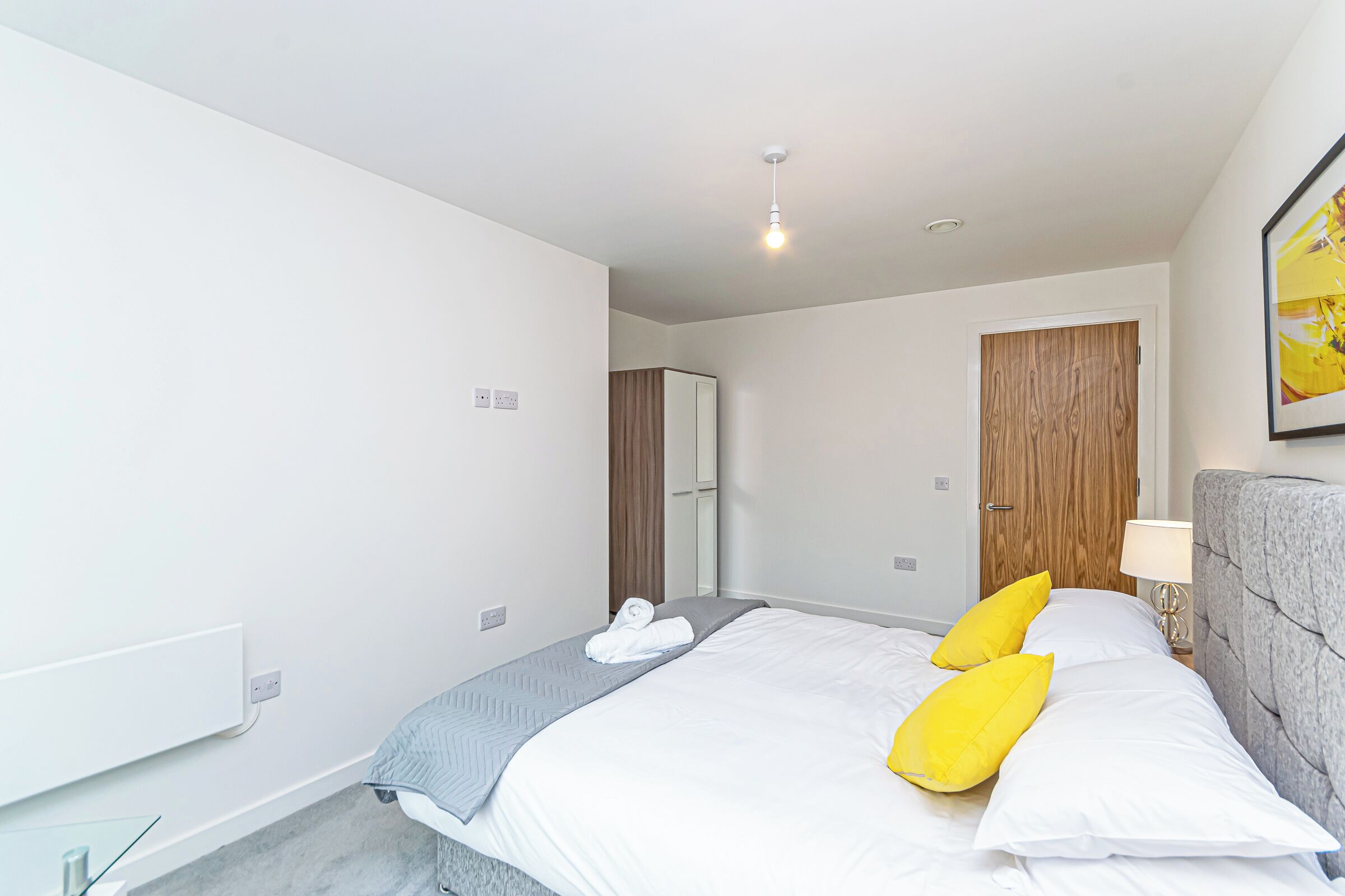A modern hotel room with a neatly made bed, a wooden door, and a bedside table with a lamp.