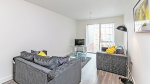 Een moderne woonkamer met een grijze bank, een kleinere grijze fauteuil, een glazen salontafel en een flatscreen televisie.