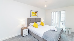 Een moderne slaapkamer met een grijs gestoffeerd bed, witte beddengoed en twee gele kussens. Er staat een nachtkastje met een lamp, een klein wit ladeblok, en een ingelijste foto boven het bed. Er is een glazen tafel naast het raam, dat verticale jaloezieën heeft.