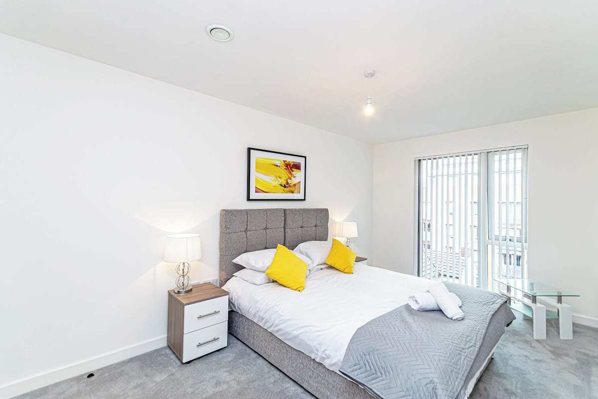 A modern bedroom with a grey upholstered bed, white bedding, and two yellow pillows. There is a bedside table with a lamp, a small white drawer, and a framed picture above the bed. A glass table is next to the window, which has vertical blinds.