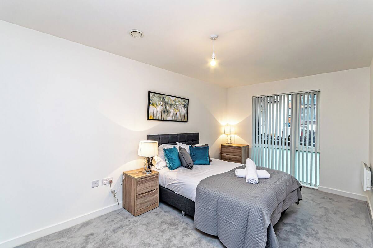 A bedroom with a bed, nightstands, a window with blinds, a painting on the wall, and a ceiling light.