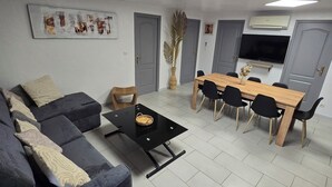 A living room with a sofa, a coffee table, a dining table with chairs, a flat-screen TV mounted on the wall, and a large window. - Family getaway near the sea (Sainte-Marie-la-Mer)