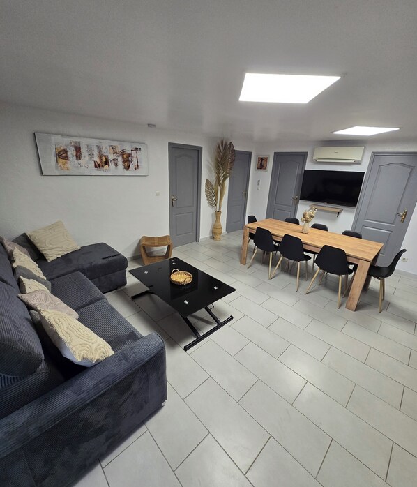 A living room with a sofa, a coffee table, a dining table with chairs, a flat-screen TV mounted on the wall, and a large window. - Family getaway near the sea (Sainte-Marie-la-Mer)