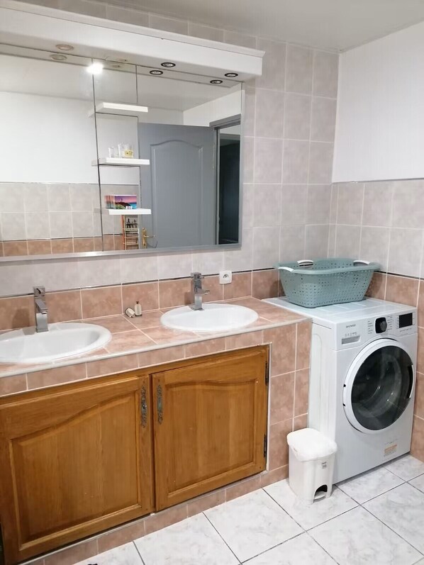 Un baño con dos lavamanos, un espejo, una lavadora y un cubo de basura pequeño.