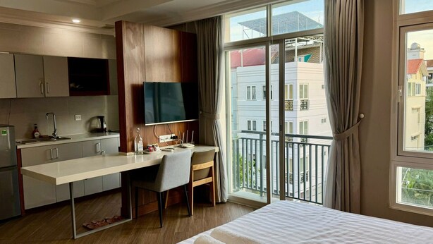 A hotel room with a bed, a desk with a chair, a TV, a kitchenette, and a balcony with curtains.