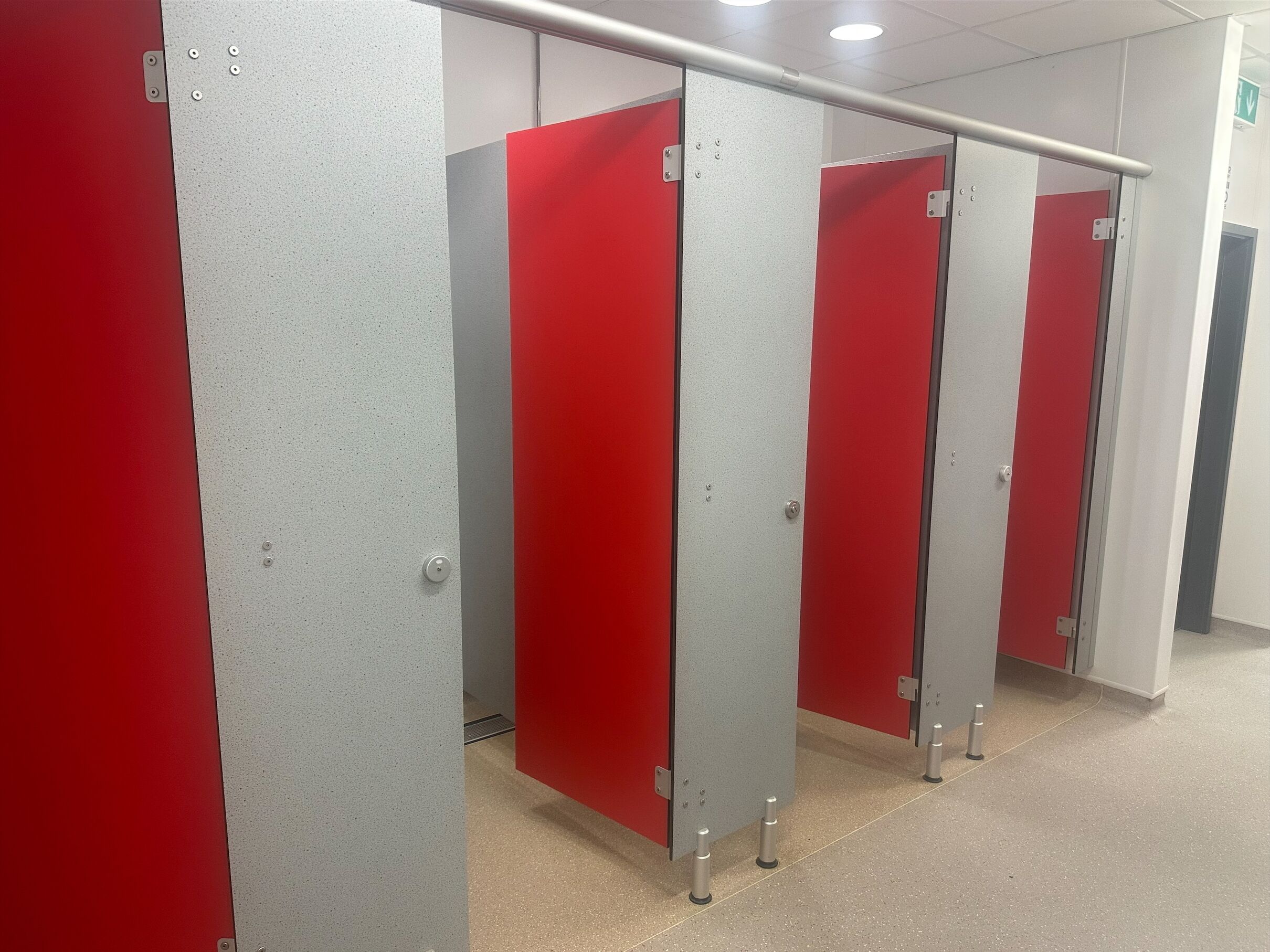A row of red and grey partitioned restroom stalls with a metal handrail above.