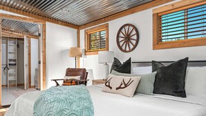 Une chambre à coucher avec un grand lit, un plafond en bois, une fenêtre avec des stores, une horloge fixée au mur et une porte donnant sur une salle de bain.