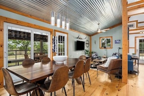 A spacious dining area with a large wooden table and chairs, a TV mounted on the wall, and a rustic ceiling.