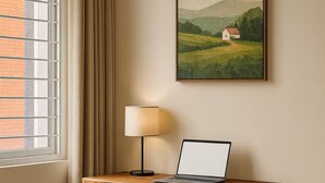 Escrivaninha no canto com um laptop, uma lâmpada e uma cadeira em um quarto com janela e um quadro na parede.
