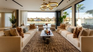A spacious living room with a large window offering a view of a beach and resort buildings. There are two sofas, a coffee table, and a chandelier. - Villa Sarai by Luxury Explorers Collection (Dubai)