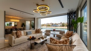 A modern living room with a sofa, coffee table, and chairs, a dining area, and a large window with a view of a pool and houses. - Villa Sarai by Luxury Explorers Collection (Dubai)