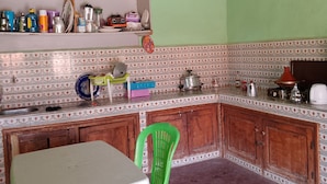 Una cocina con pared verde, techo de madera y salpicadero de azulejos. Hay una mesa y una silla verde, y varios utensilios de cocina en estantes y encimeras.