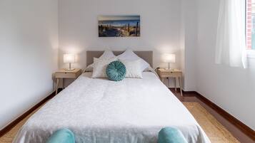 Una cama bien hecha con ropa blanca y una sola almohada de color aguamarina, flanqueada por dos mesitas de noche con lámparas, en una habitación con pared blanca lisa y una ventana con cortinas.