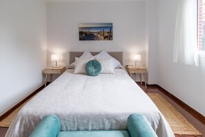 Un lit bien fait, avec une literie blanche et un seul coussin d’accent de couleur turquoise, flanqué de deux tables de chevet avec des lampes, dans une pièce dont le mur est d’un blanc uni et qui possède une fenêtre avec des rideaux.