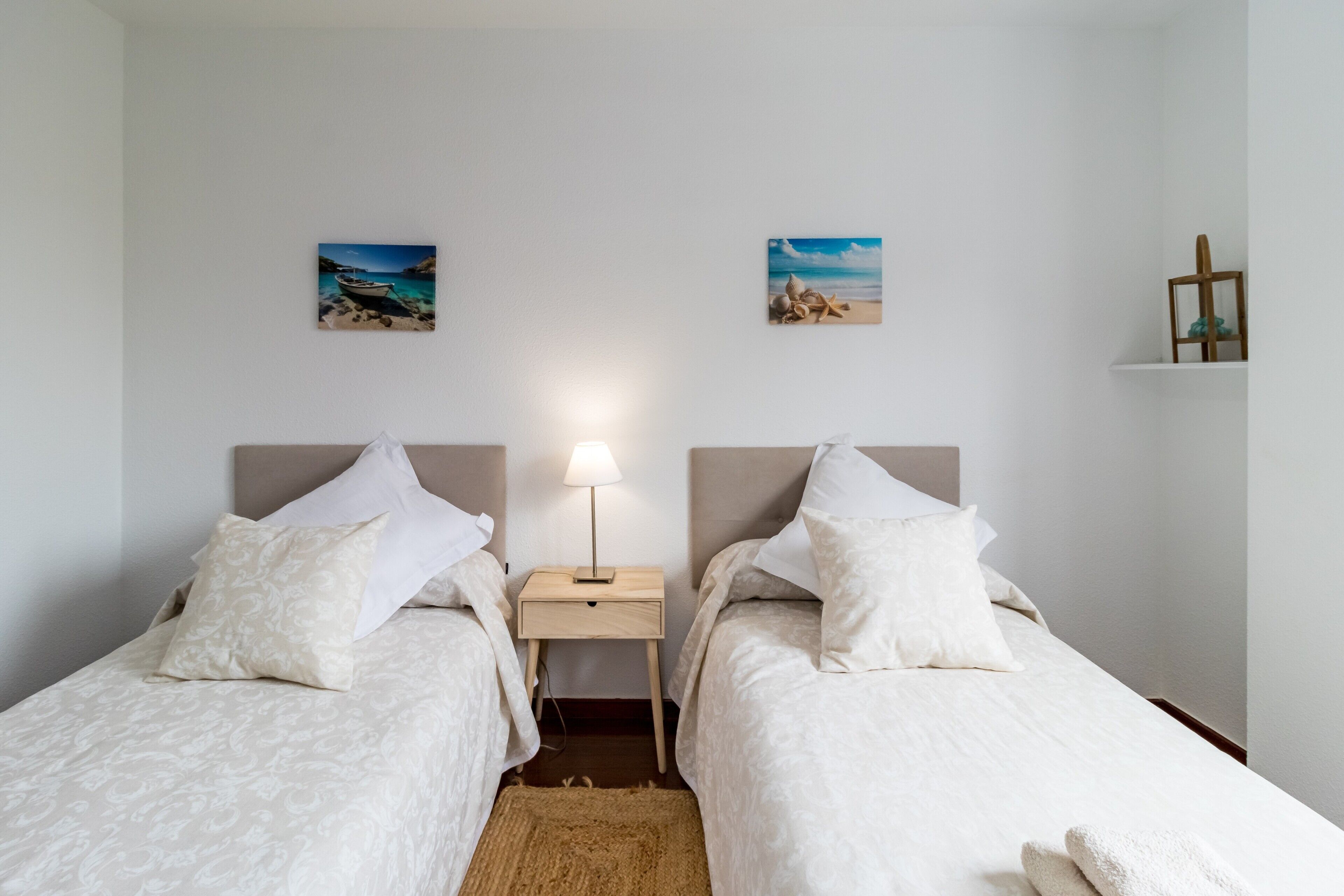 Een slaapkamer met twee bedden, een nachtkastje met een lamp en wanddecoratie met een strandtafereel.