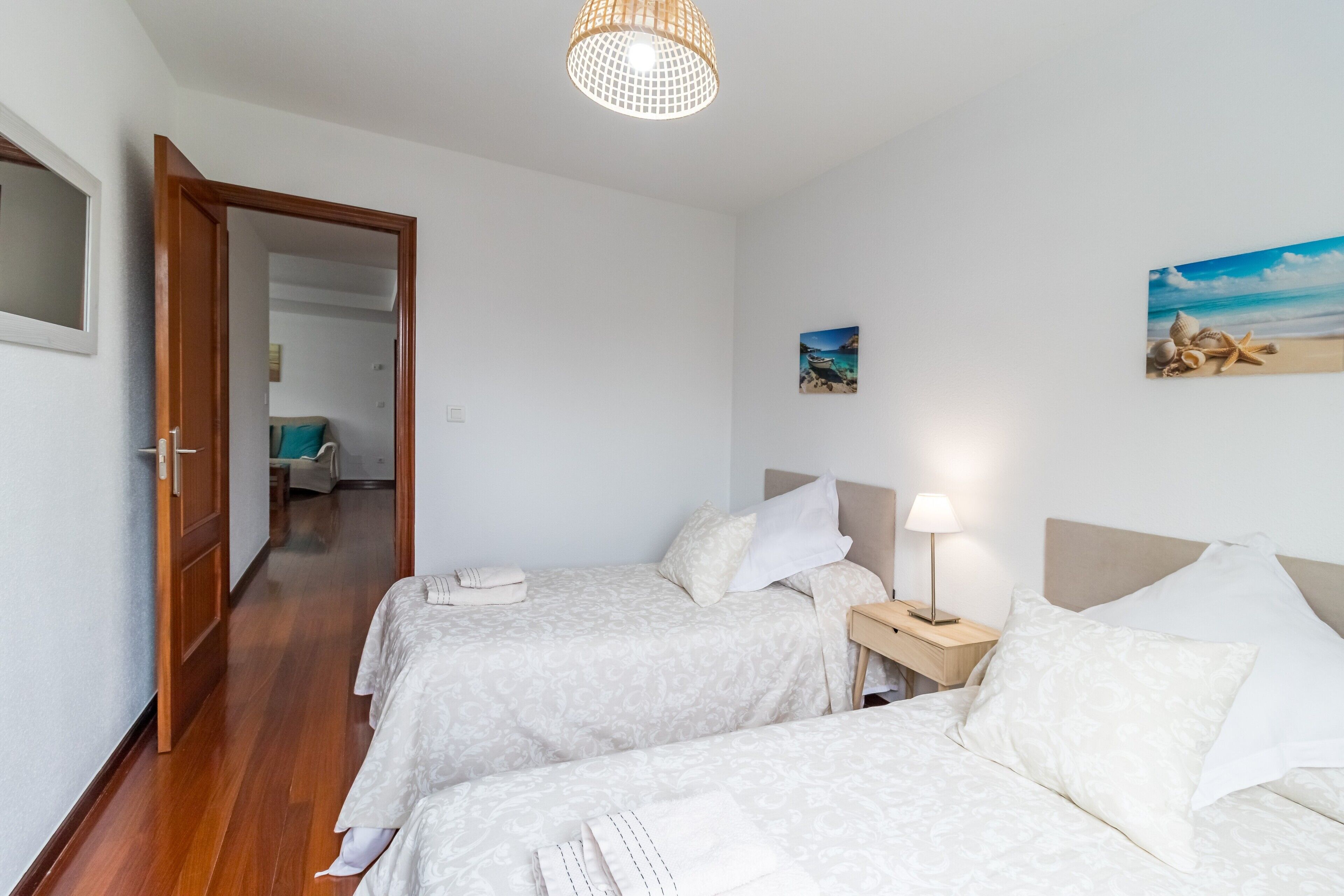 Een hotelkamer met twee bedden, een houten vloer, een deur naar een andere kamer, een tafeltje met een lamp en een schilderij met een strandthema aan de muur.