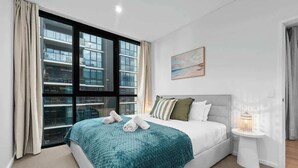 A modern bedroom with a large bed, a view of a building through a window, a bedside table with a lamp, and a painting on the wall.