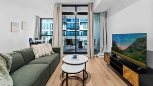 Una sala de estar moderna con un sofá verde, una mesita blanca, un televisor sobre un mueble de madera y un balcón con puertas de cristal.