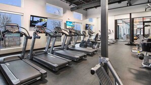 Een sportschool met loopbanden langs een muur met grote ramen, een televisiescherm en fitnessapparatuur op de achtergrond.