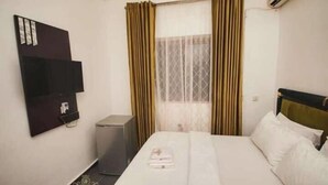 A hotel room with a bed, a TV mounted on the wall, a small desk, and a window with curtains. - Crystalem Luxury Hotel (Uyo)