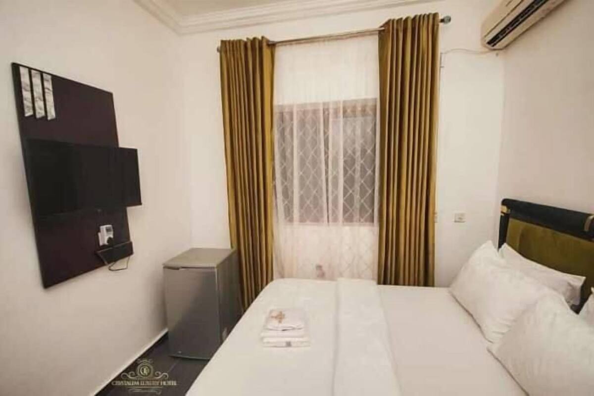 A hotel room with a bed, a TV mounted on the wall, a small desk, and a window with curtains.
