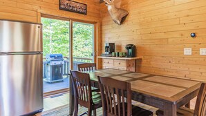 A wooden cabin interior with a dining table, chairs, a refrigerator, a mounted deer head, a coffee maker, and a grill outside.