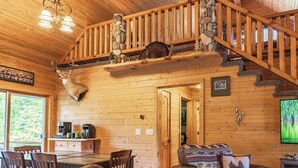 A wooden cabin interior with a dining area, a staircase with a stone wall, and a mounted deer head.