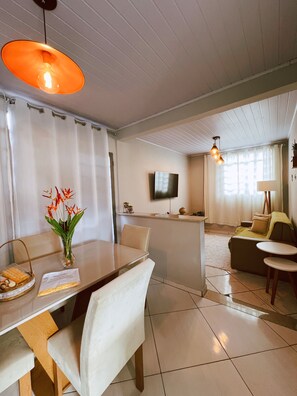Una habitación de hotel moderna con zona de comedor, sofá, televisor y ventana con cortinas.