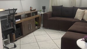 Een moderne woonkamer met een flatscreen televisie, een bruine bank, een klein rond tafeltje en een betegelde vloer.