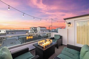 A rooftop terrace with a fire feature, outdoor seating, and string lights against a sunset sky. - Ocean Views, Steps to Beach, Rooftop, Firepit (San Diego)