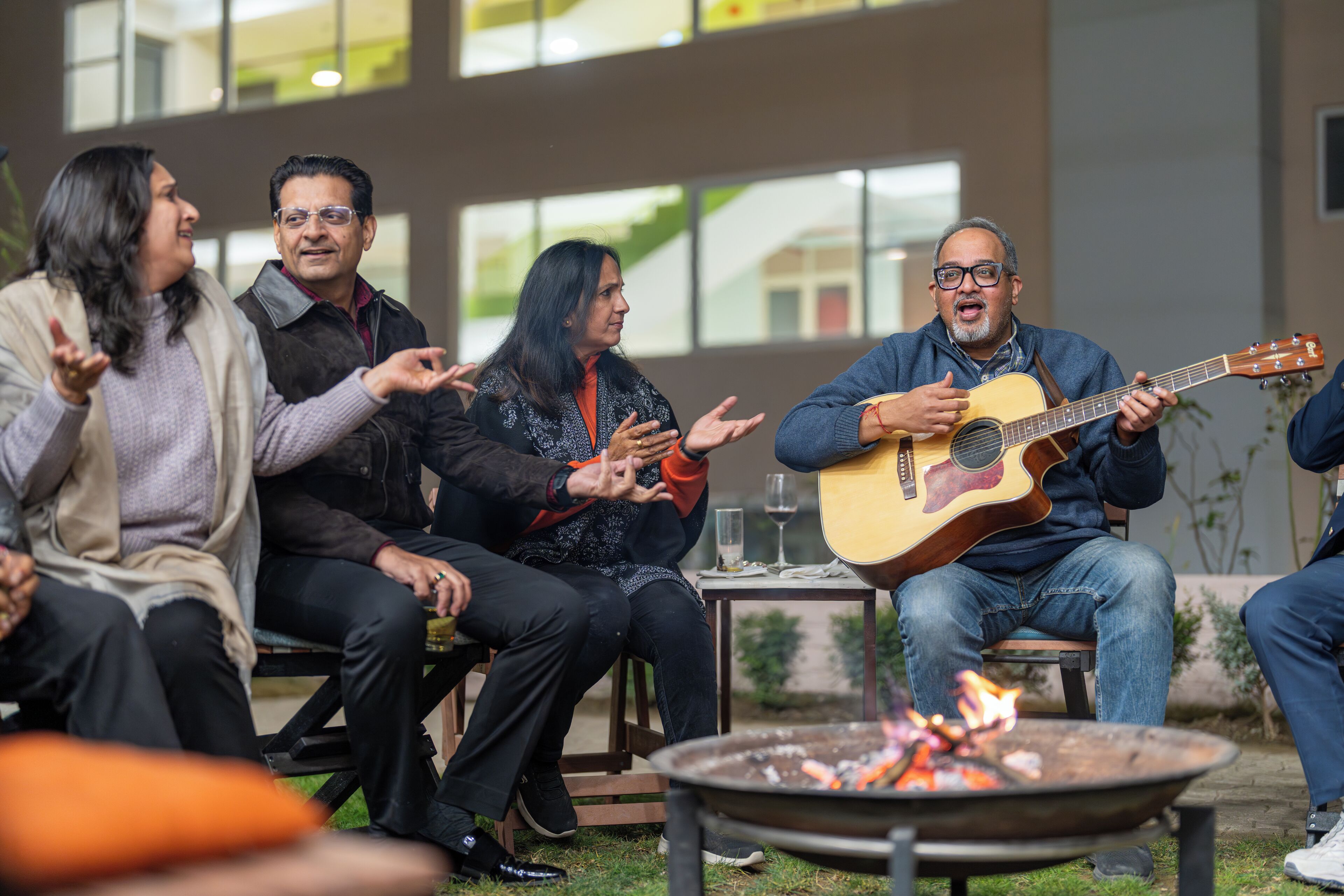 A group of people sitting outdoors around a fire pit, with a building in the background.