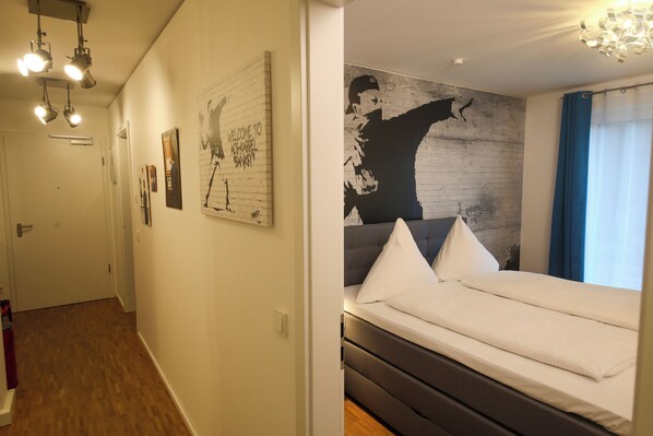 A hotel room with a bed, a wall with a large black and white mural, a hallway with wooden flooring, and a door.