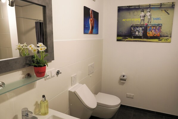 A bathroom with a toilet, sink, and mirror. There is a potted plant on the sink counter and a poster on the wall.