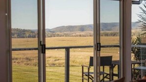 A room with a large sliding glass door leading to a balcony with a view of a field and hills. There are two chairs and a small table on the balcony.