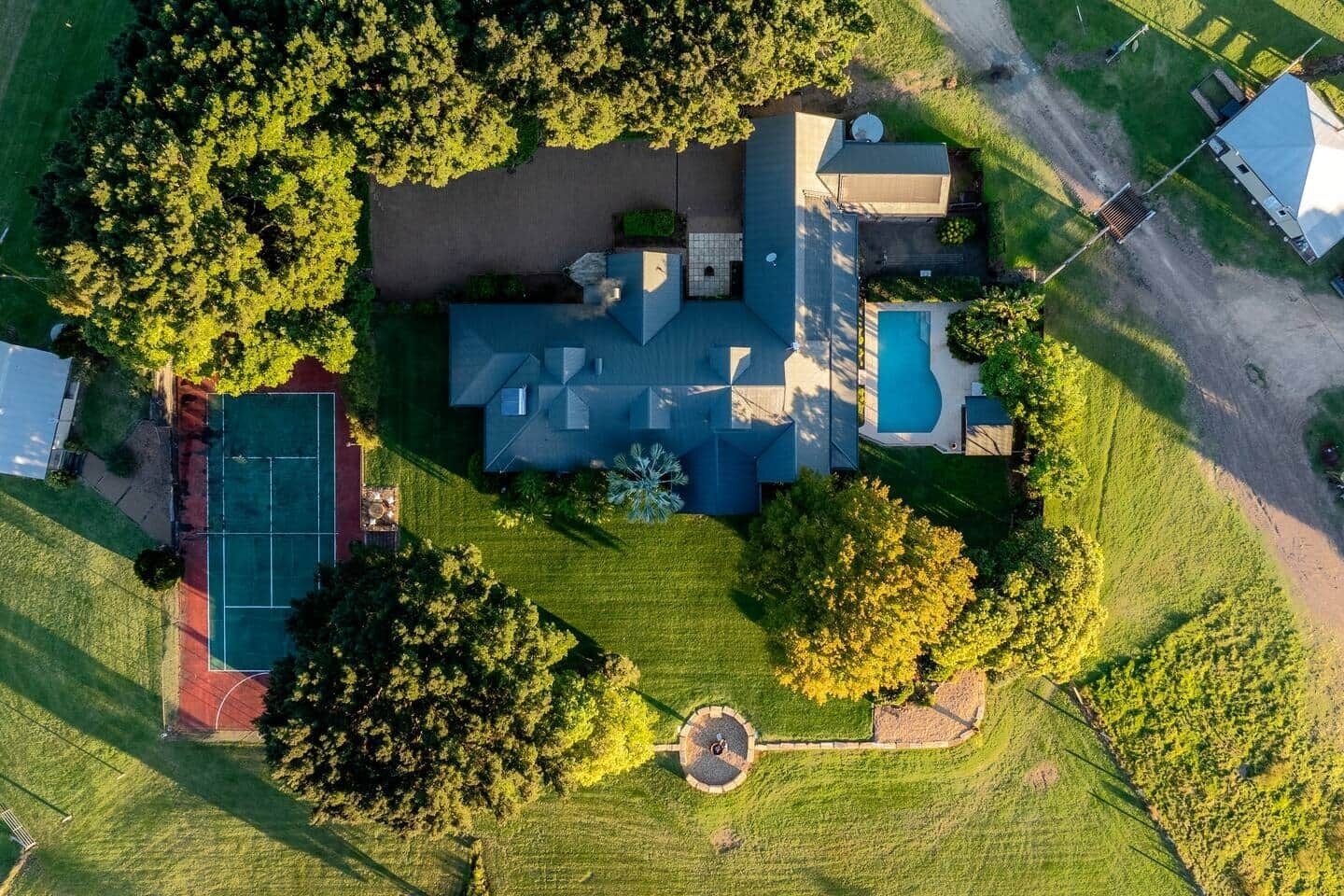 An aerial view of a residential property with a large house, a swimming pool, a tennis court, and surrounding greenery.