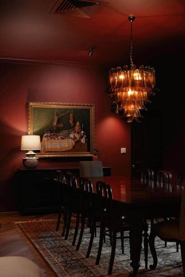 A dining room with a large wooden table, dark chairs, a chandelier, a painting on the wall, and a lamp on a side table.