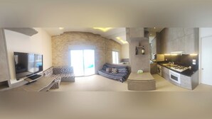 A modern living room with a flat-screen TV, a sofa, a coffee table, and a kitchen area with a gas stove and cabinets.