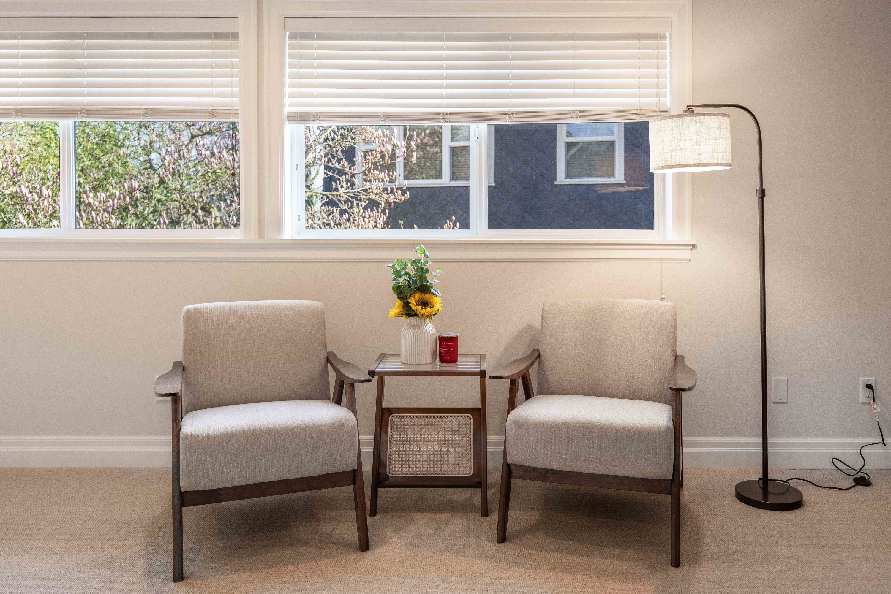 A room with two chairs, a small table with a vase and a red can, a floor lamp, and a window with blinds.