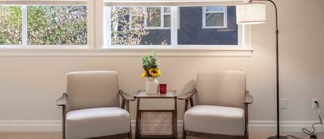 A room with two chairs, a small table with a vase and a red can, a floor lamp, and a window with blinds.