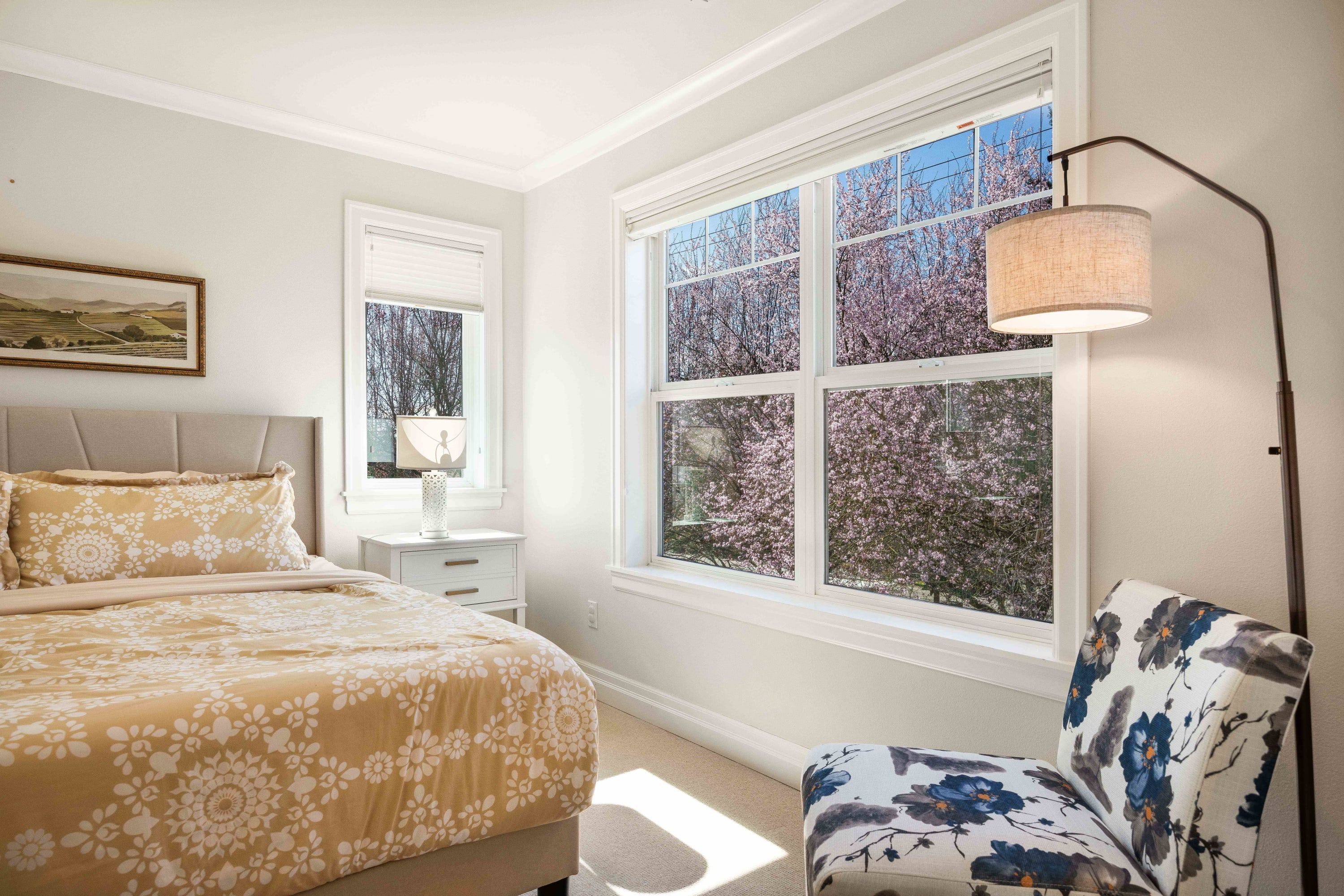 A bedroom with a large window, a bed with floral bedding, a chair with a floral pattern, and a bedside table with a lamp.