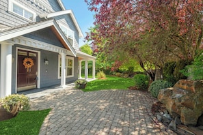 A residential house with a covered porch, a front door with a wreath, and a well-maintained garden with a tree and shrubs.