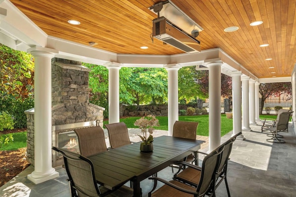 An outdoor patio with a stone fireplace, a dining table with chairs, and a pergola structure.